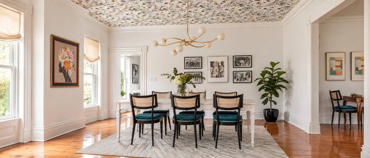 The dining room after: a botanical print across the ceiling, a branching brass chandelier with opal globes, cane-back chairs with teal velvet seats around a whitewashed table, a gallery wall and a fiddle-leaf fig.