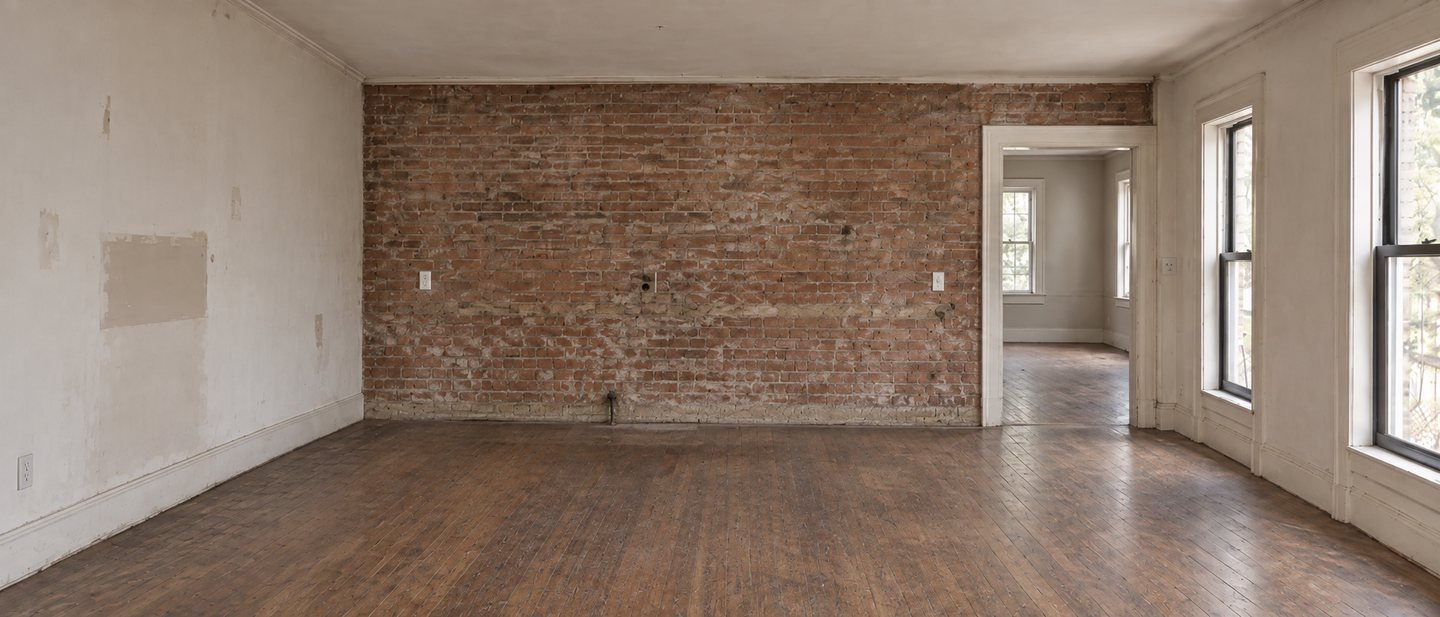 The kitchen before: an empty room with an exposed brick wall, worn pine floorboards and a doorway to the next room.