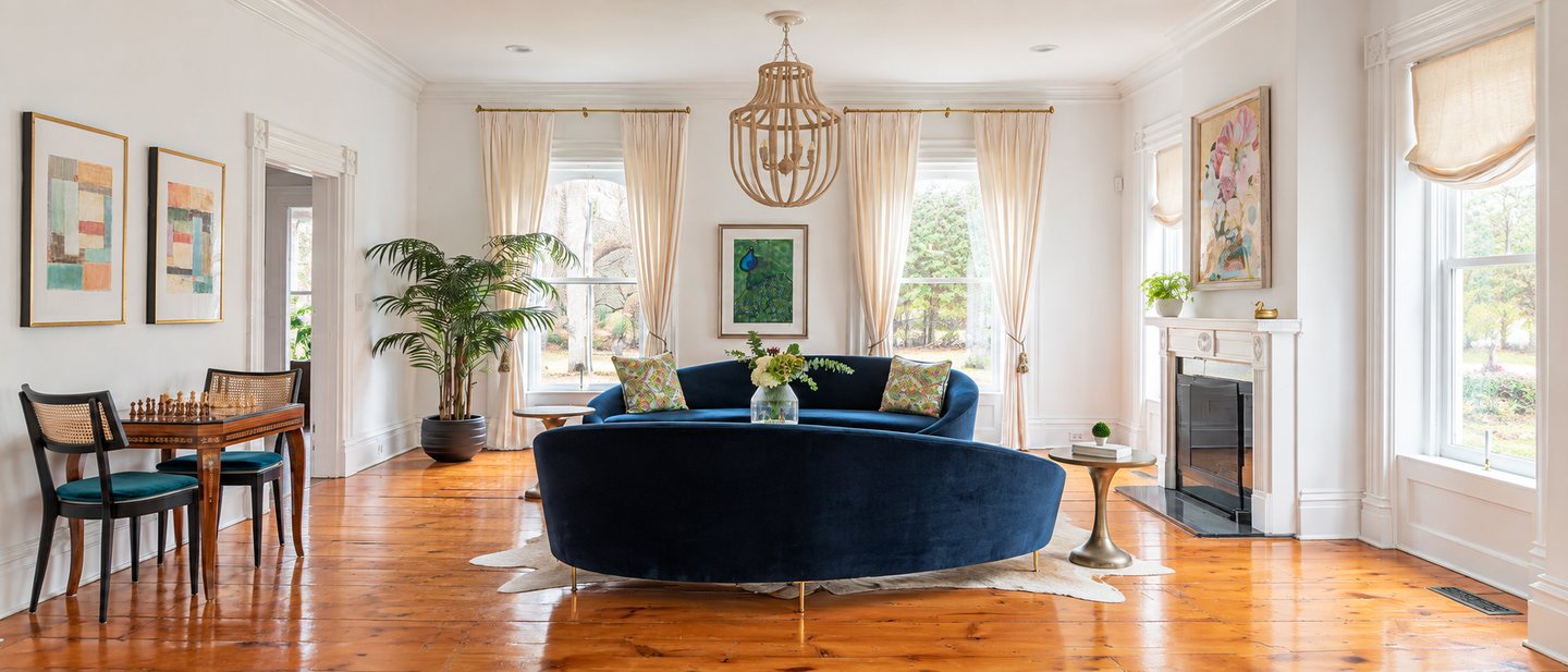 The living room after: a curved sapphire velvet sofa in the middle of the room, linen drapes, a wood-bead chandelier, a floral painting over the mantel and a chess table in the corner.