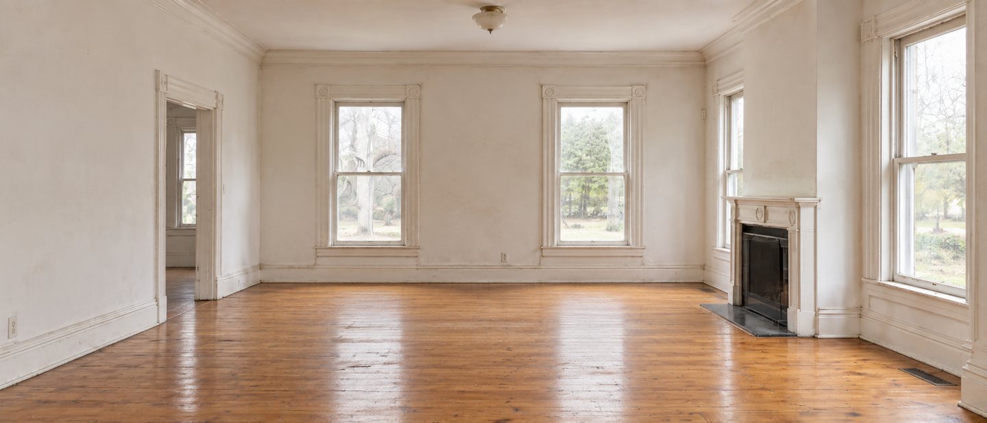 The living room before: an empty white parlour with tall sash windows, an original mantel and pine floors.